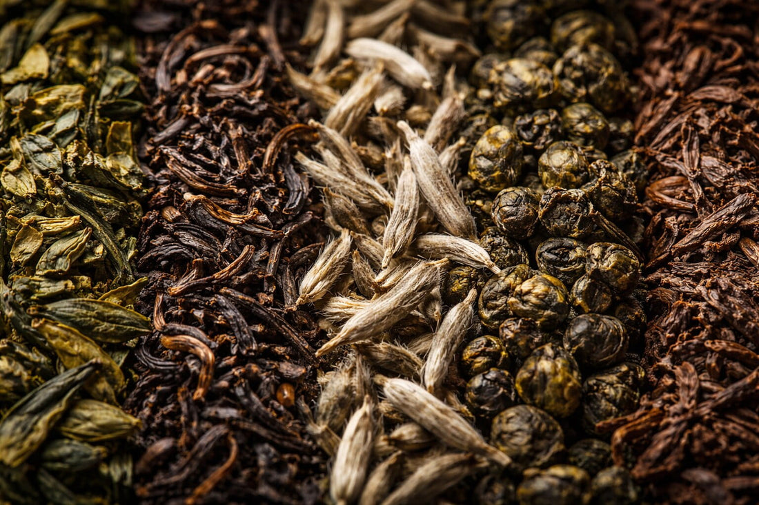 Gros plan macro de différents thés en vrac alignés côte à côte (thé vert, noir, bourgeons blancs, oolong et pu-erh), mettant en valeur leurs textures et couleurs dans une ambiance naturelle et raffinée.