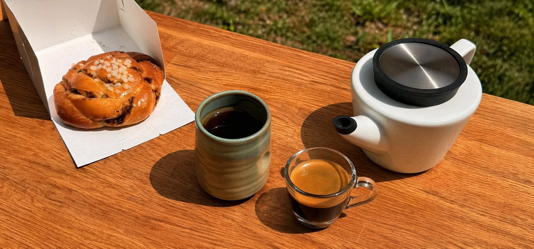 Expresso et thé servis sur une table en bois en extérieur avec une théière et une viennoiserie sous la lumière du soleil.