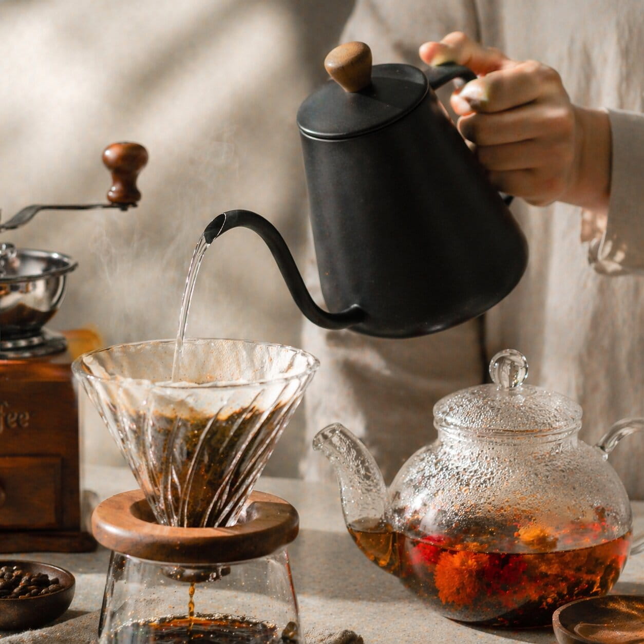 Préparation artisanale de café filtre avec verseuse et bouilloire, accompagnée d’une théière en verre contenant une infusion florale.